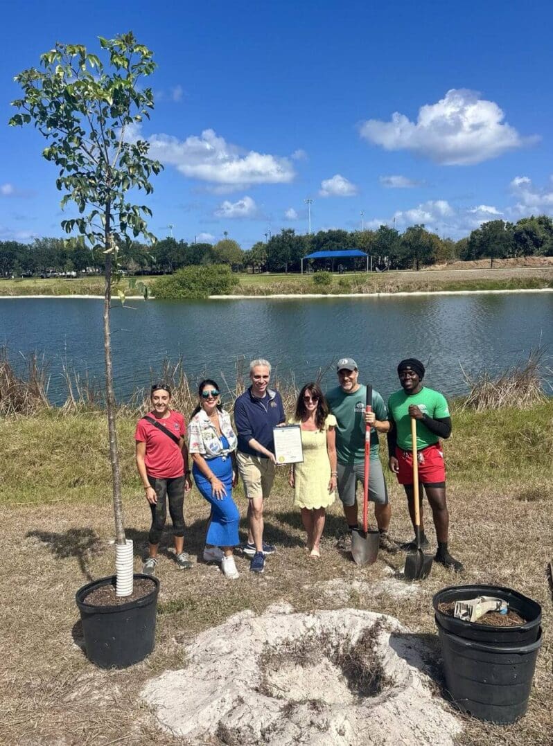 Group of people planting a tree by a lake on a sunny day.