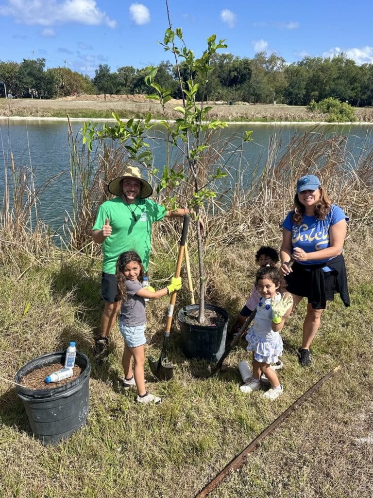 Family planting a tree together by a lakeside on a sunny day.