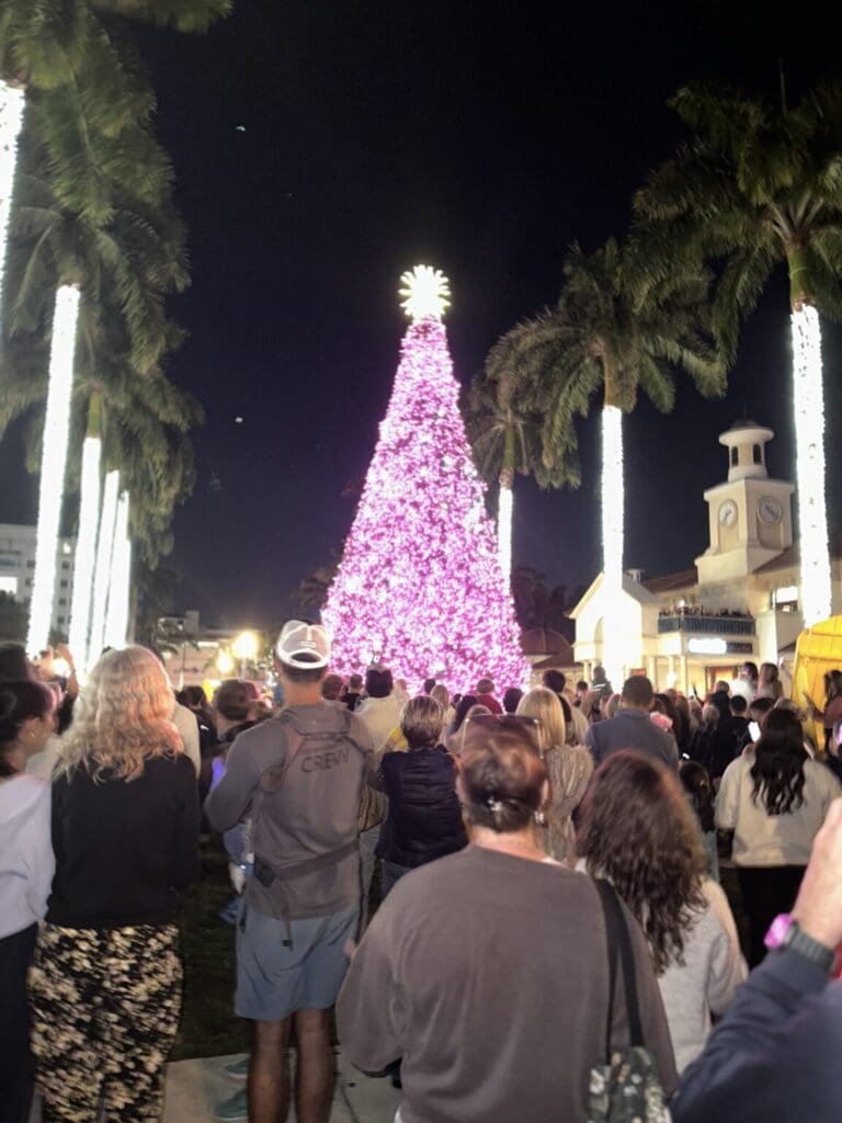 Crowd gathered around a brightly lit purple Christmas tree at night.