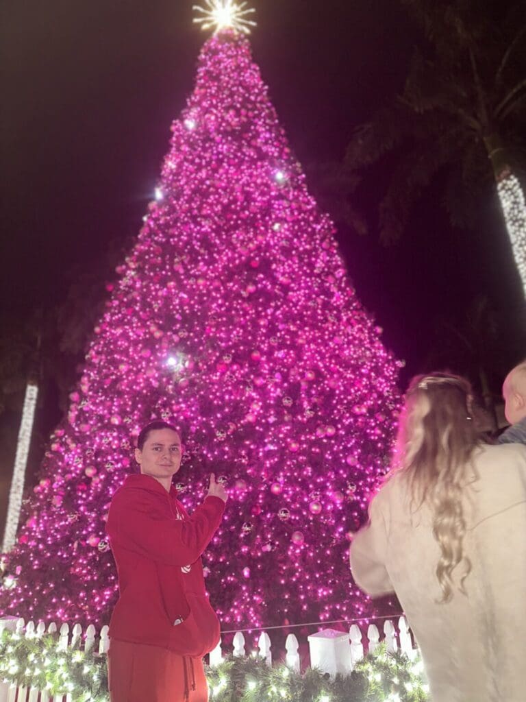 Two people posing near a large pink-lit Christmas tree at night.