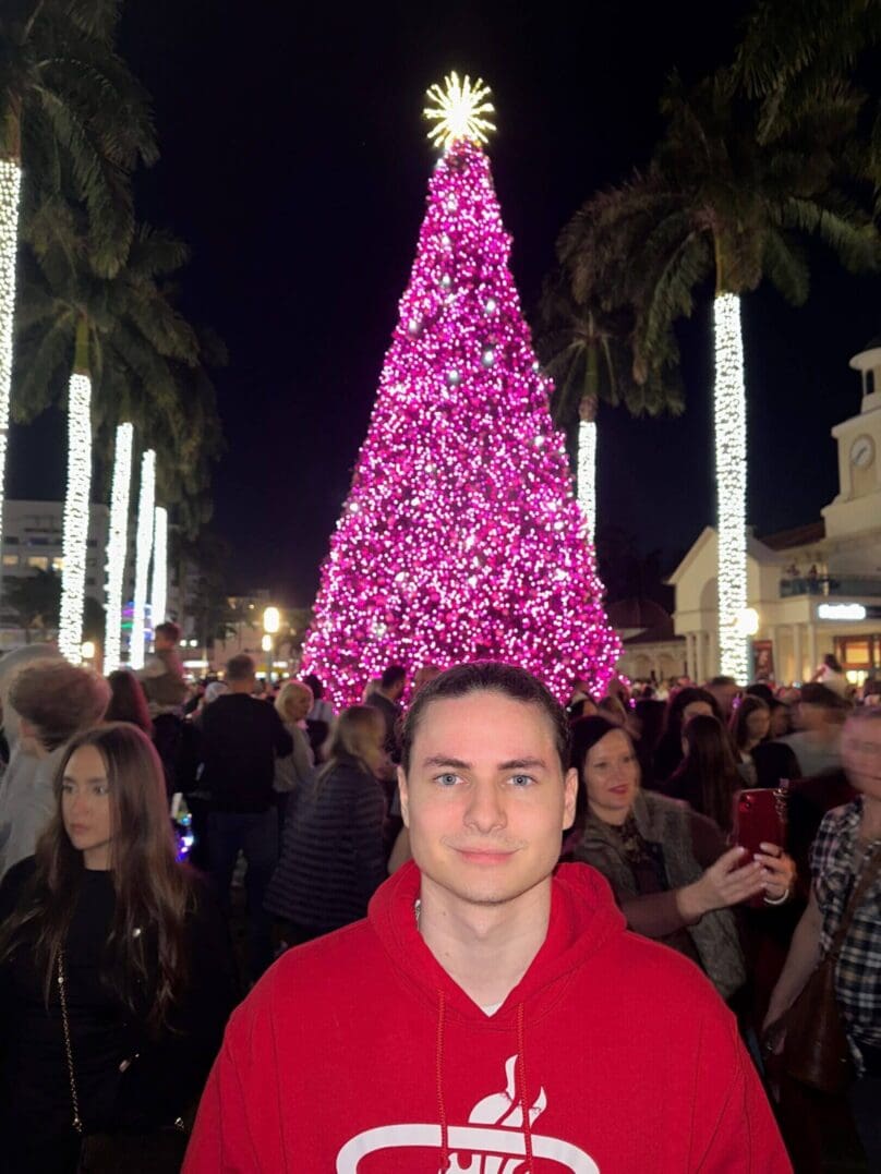 A man in a red shirt stands in front of a large pink-lit Christmas tree at night.
