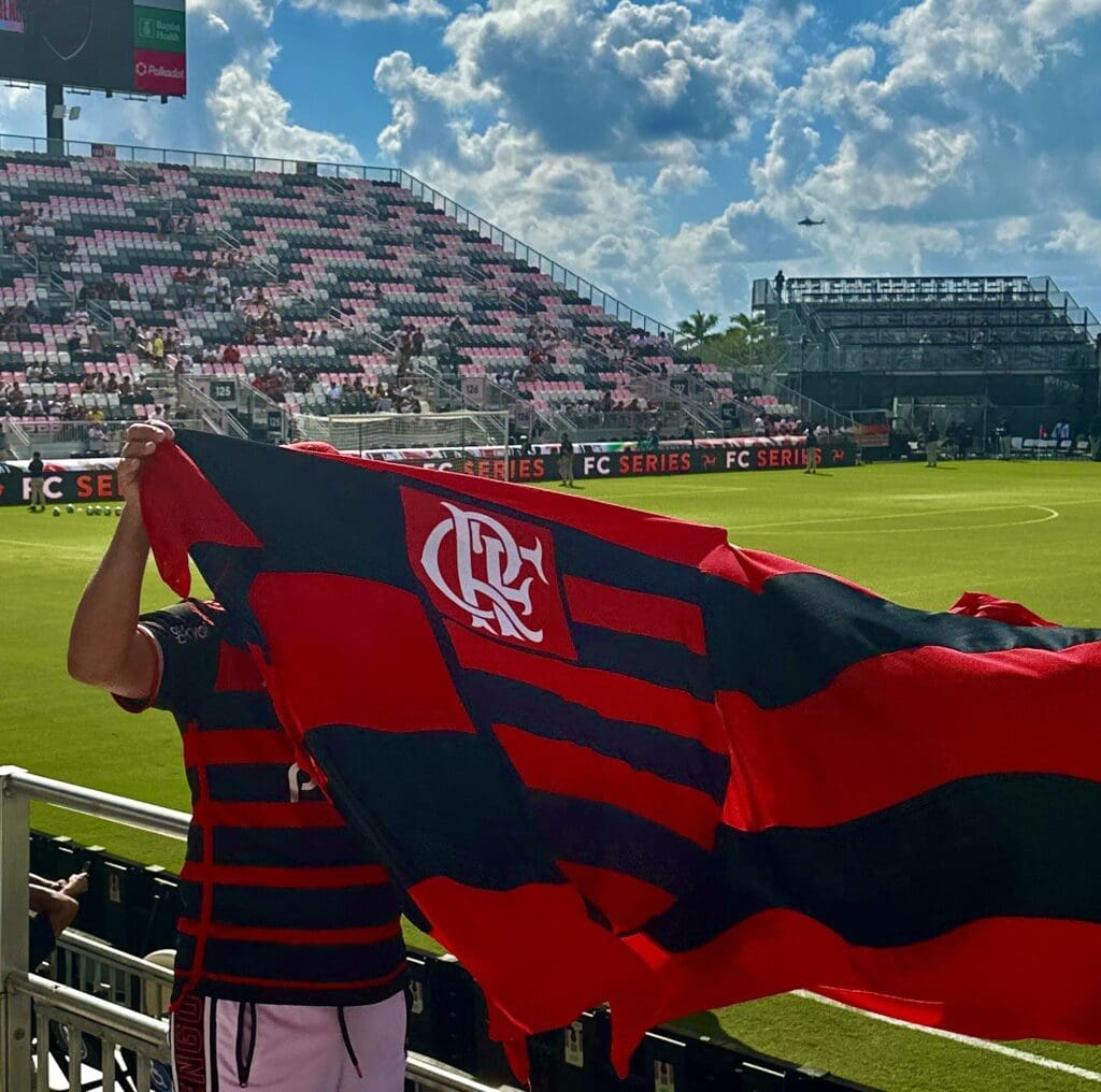 Fans waving a Flamengo flag in a stadium under a blue sky.