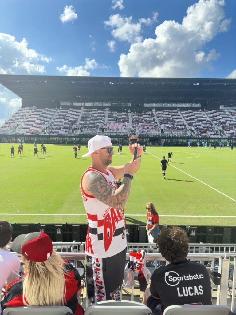 A fan taking a photo at a soccer stadium with players on the field.