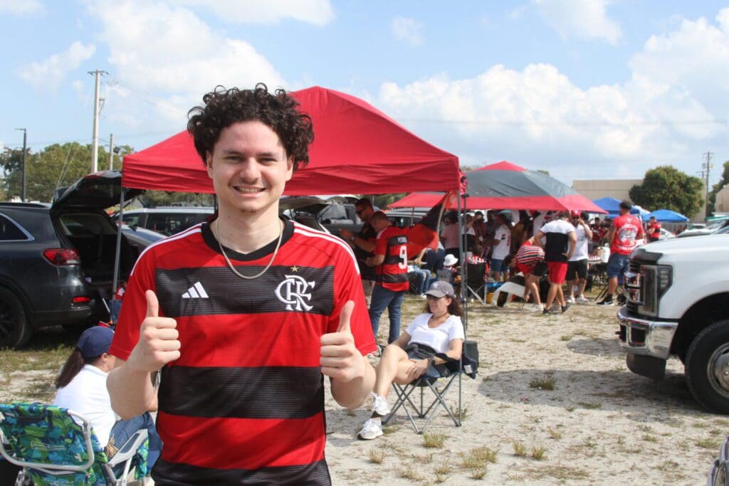 Young man in a Flamengo soccer jersey giving thumbs up at a fan event with tents and people.