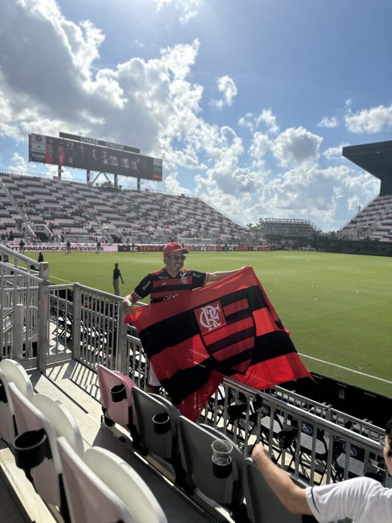 Fan waving a red and black flag at a soccer stadium under a partly cloudy sky.