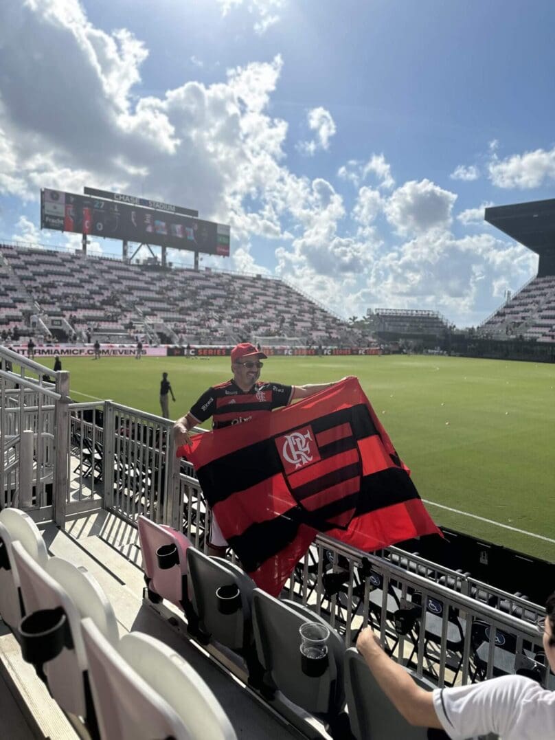 Fans wave a black and red flag at a sunny soccer stadium.