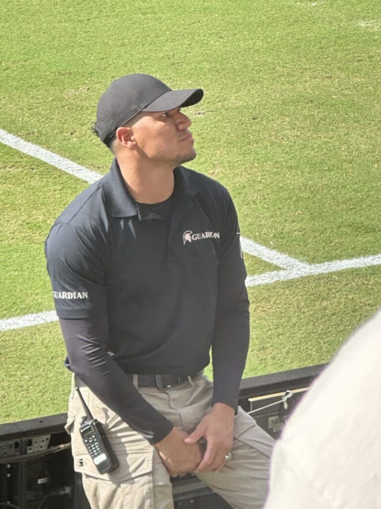 A man in a black shirt and cap sitting near a sports field.